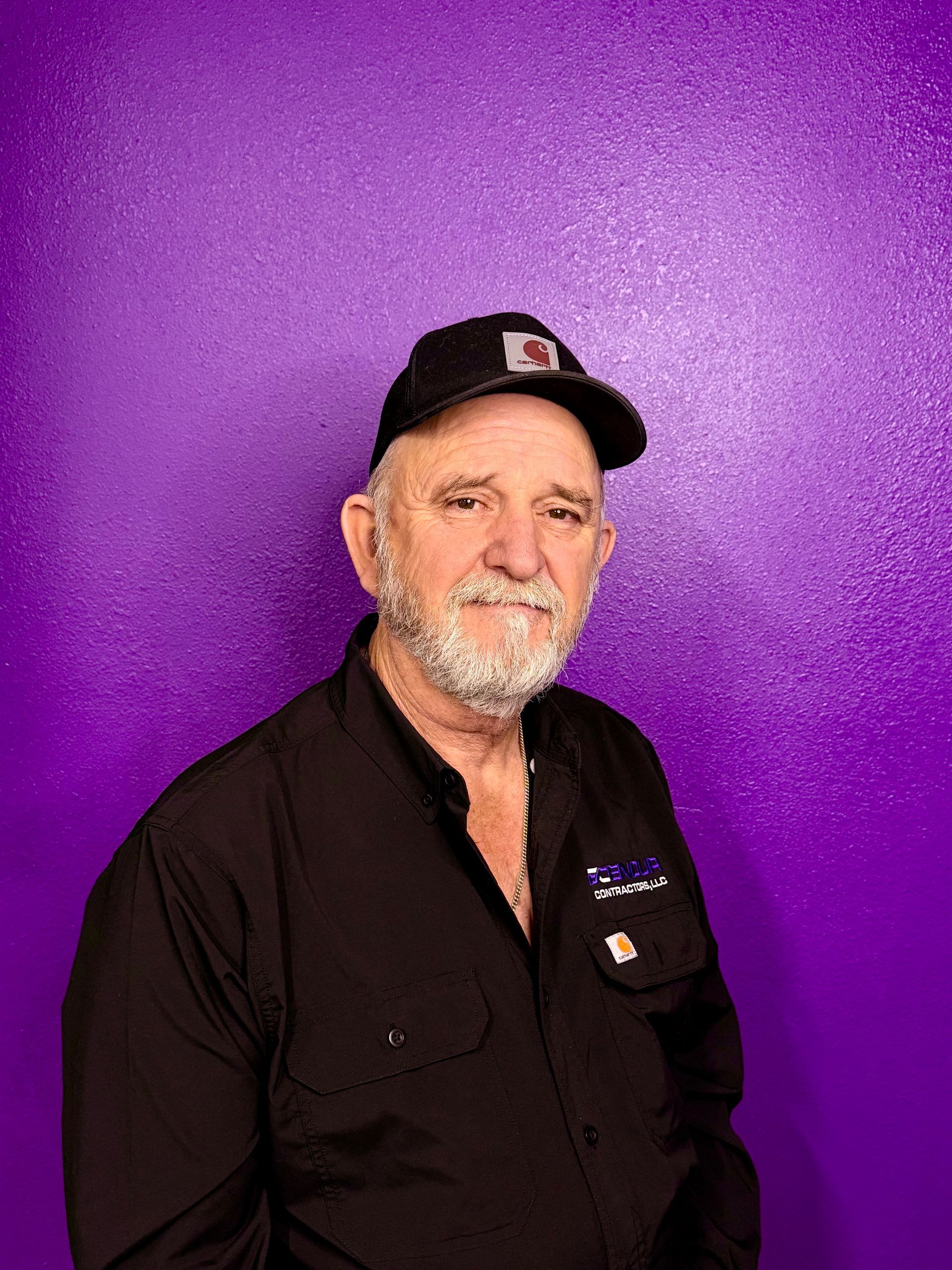 Man with a gray beard and black hat smiles against a purple background, wearing a black shirt.