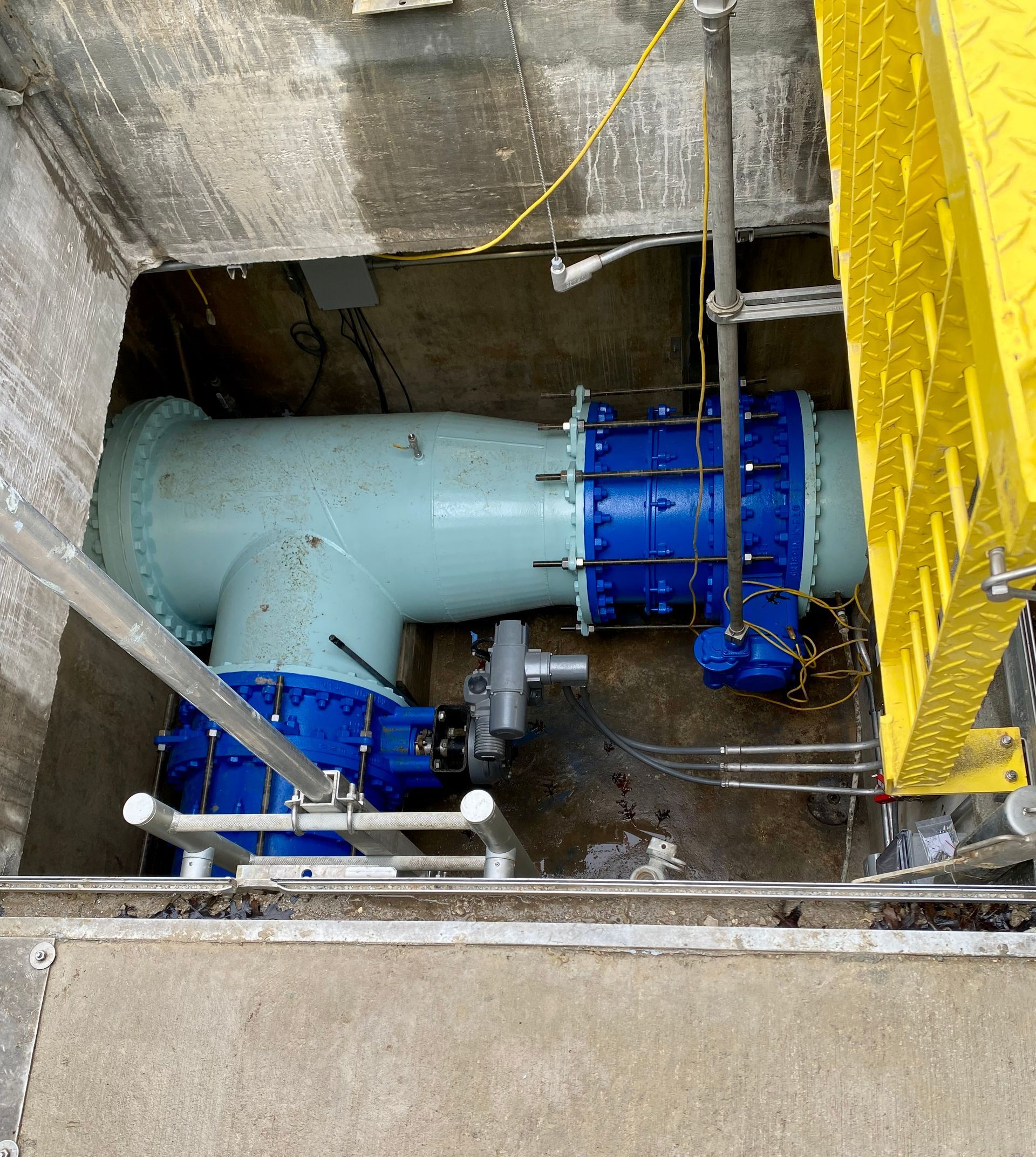 Blue industrial pipes and valves inside a concrete utility pit, with yellow safety railing.