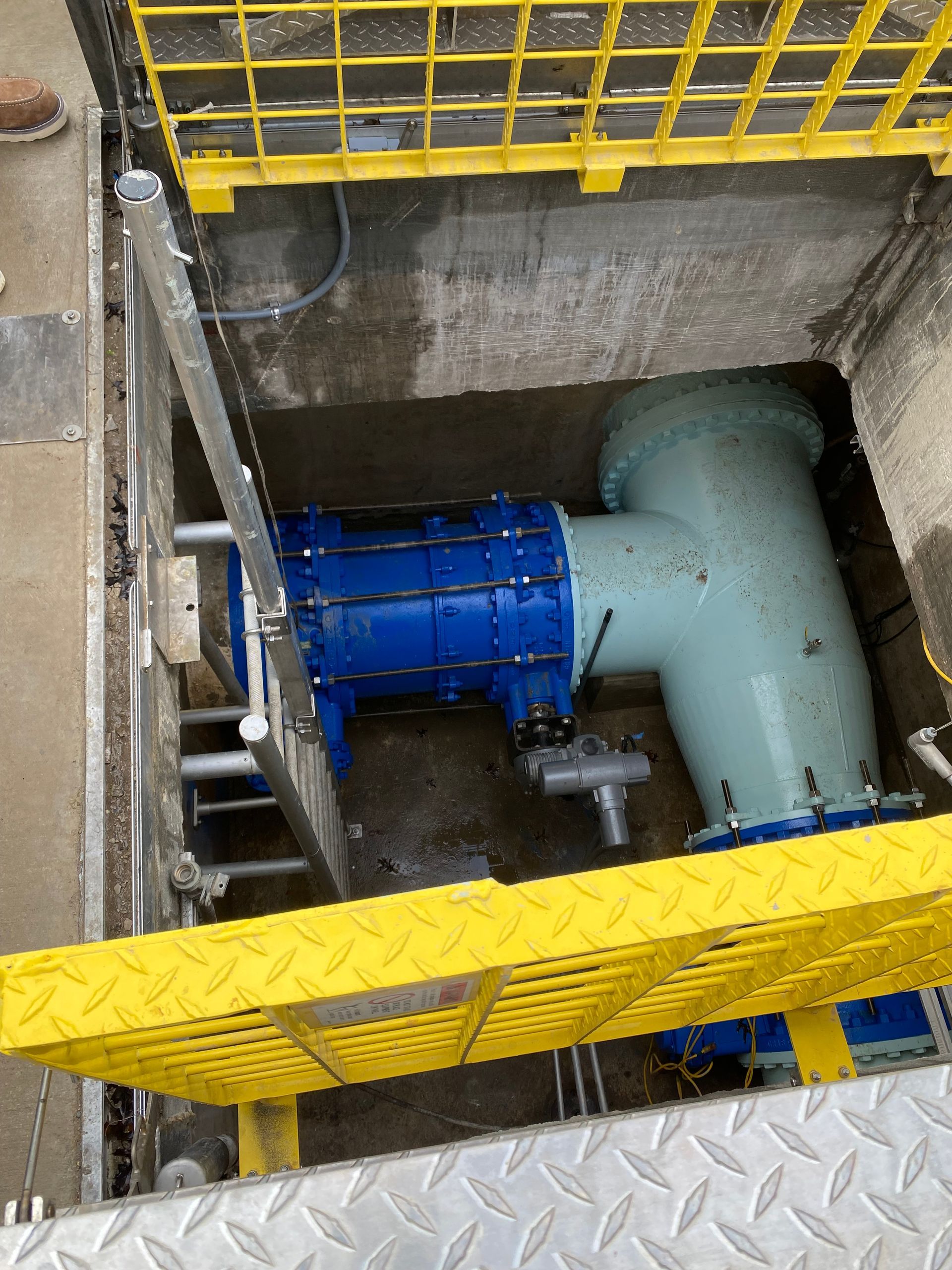 Industrial pit with blue motor and large gray pipe behind yellow safety railings and metal grating