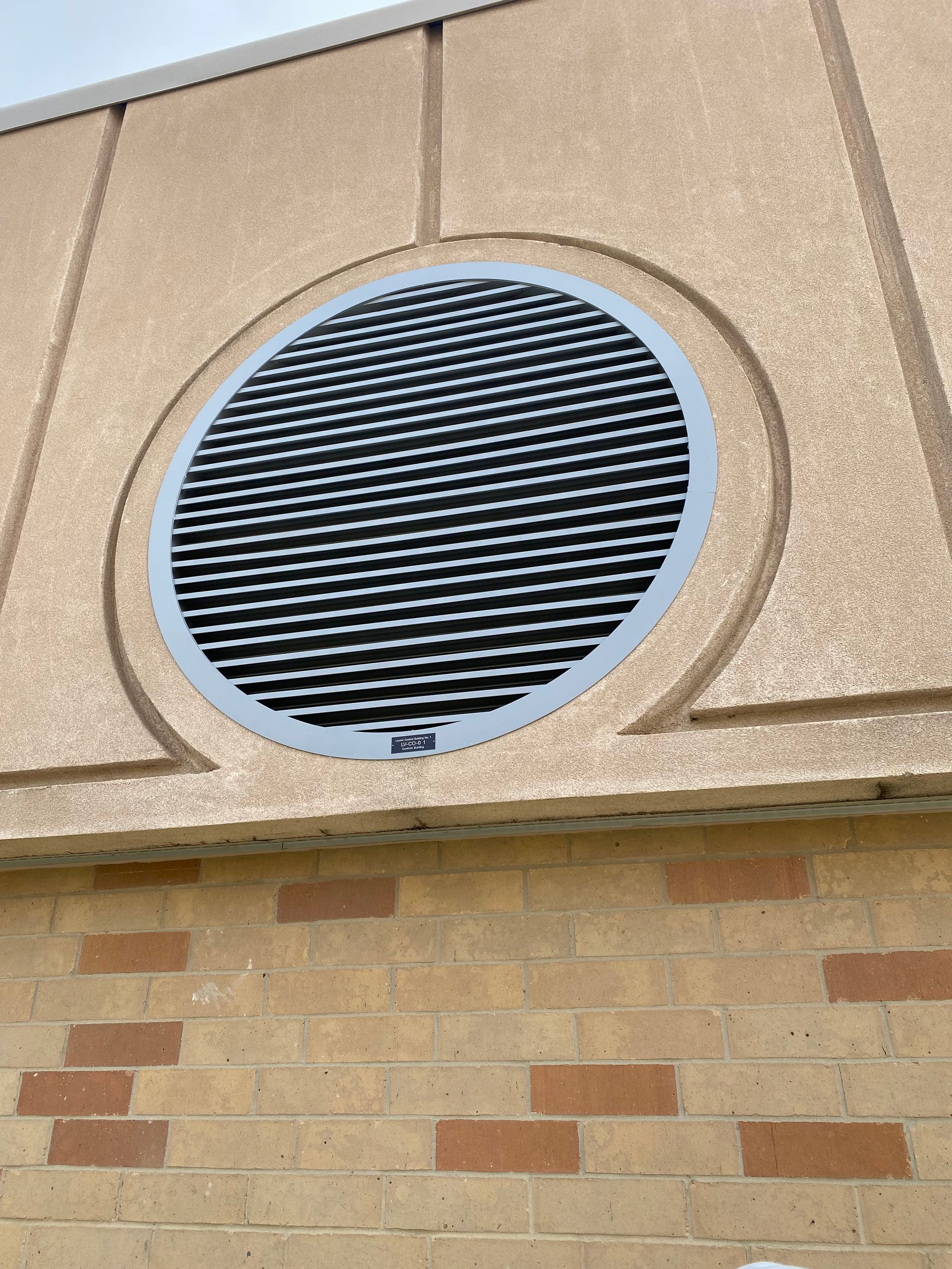 Circular gray ventilation grille on a tan building wall above brick siding