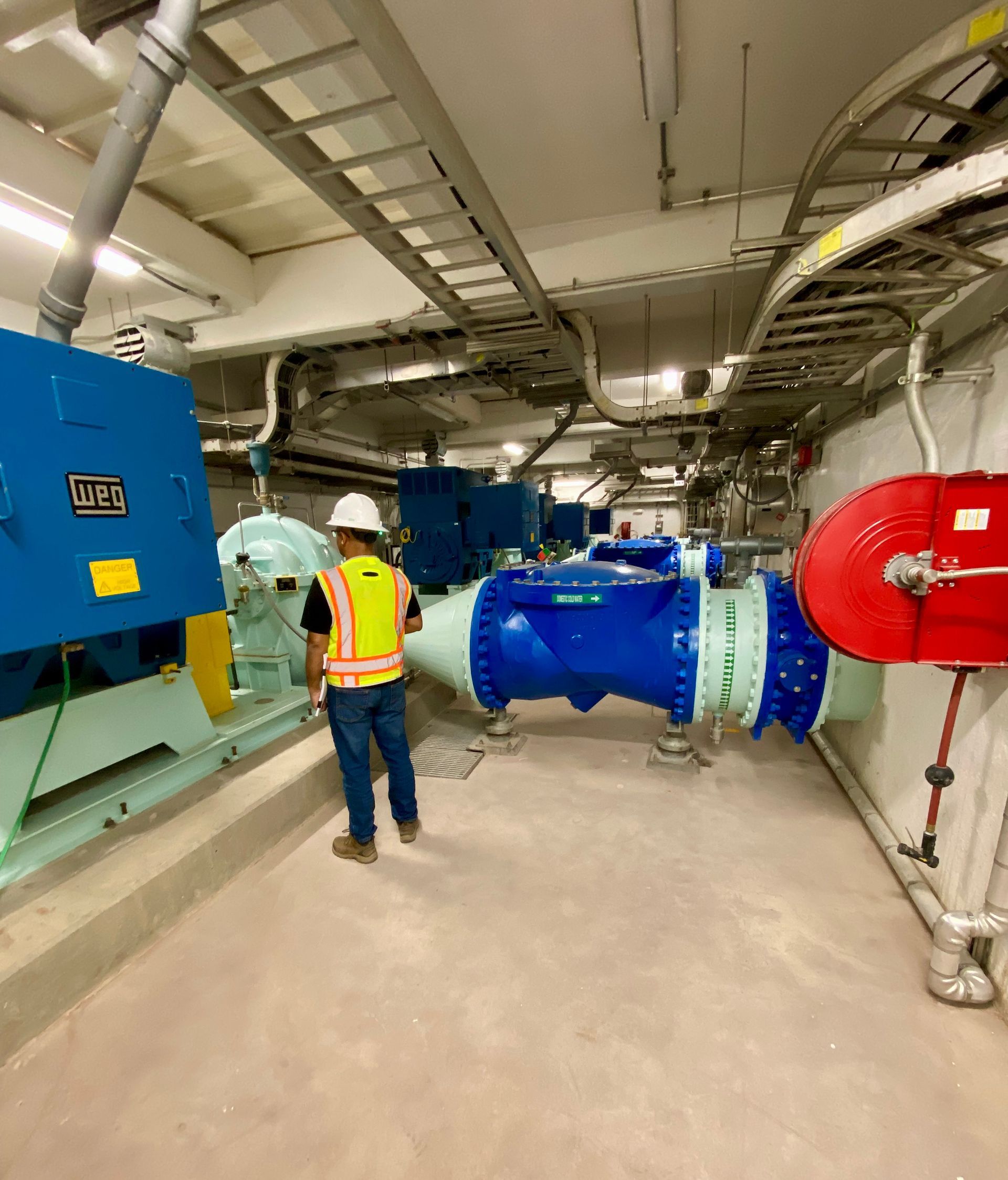 Worker in a hard hat and safety vest stands in an industrial room with large blue and red pipes and machinery.