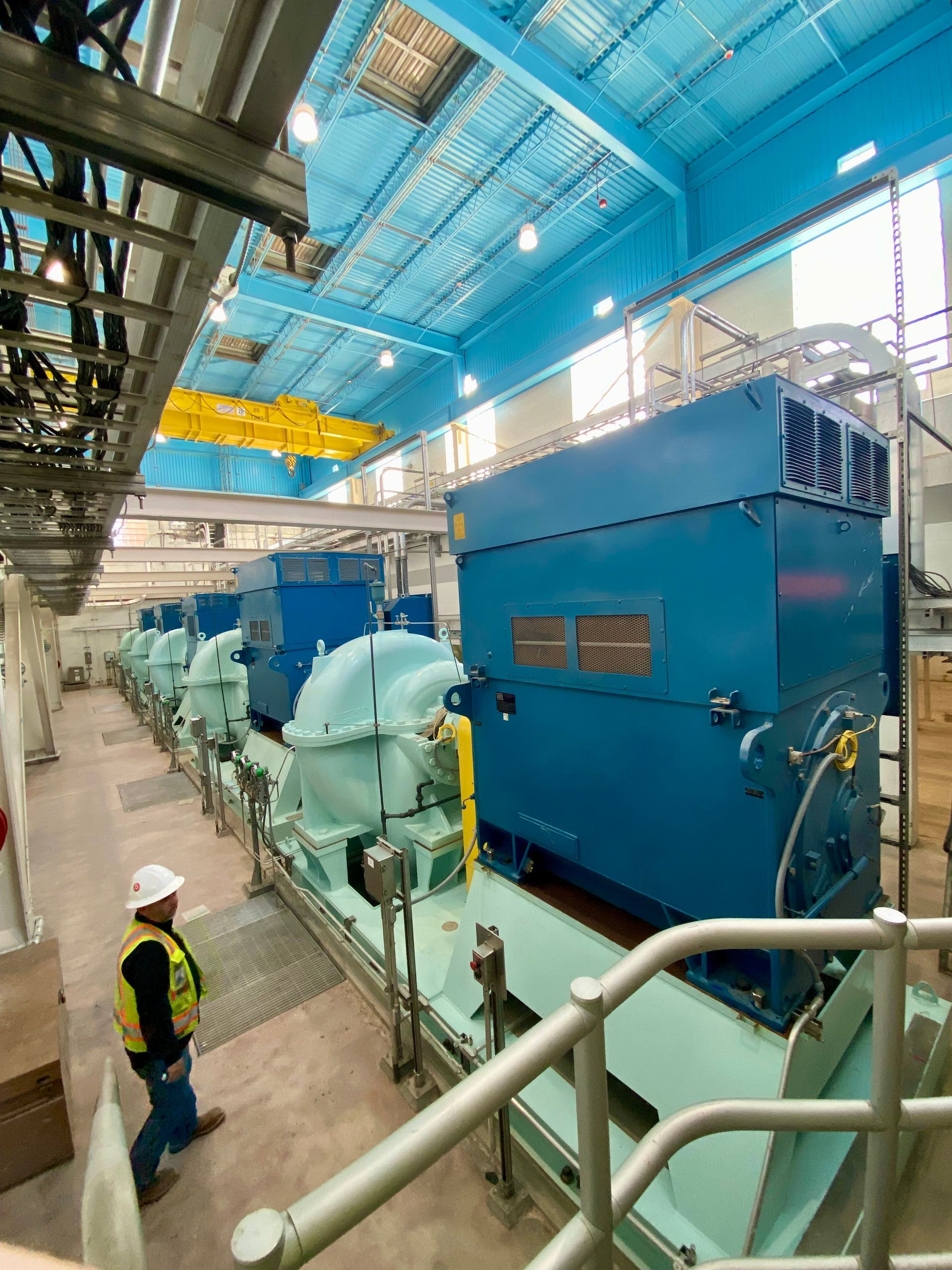 Industrial plant interior with large blue machinery and workers in safety gear under a bright skylit ceiling