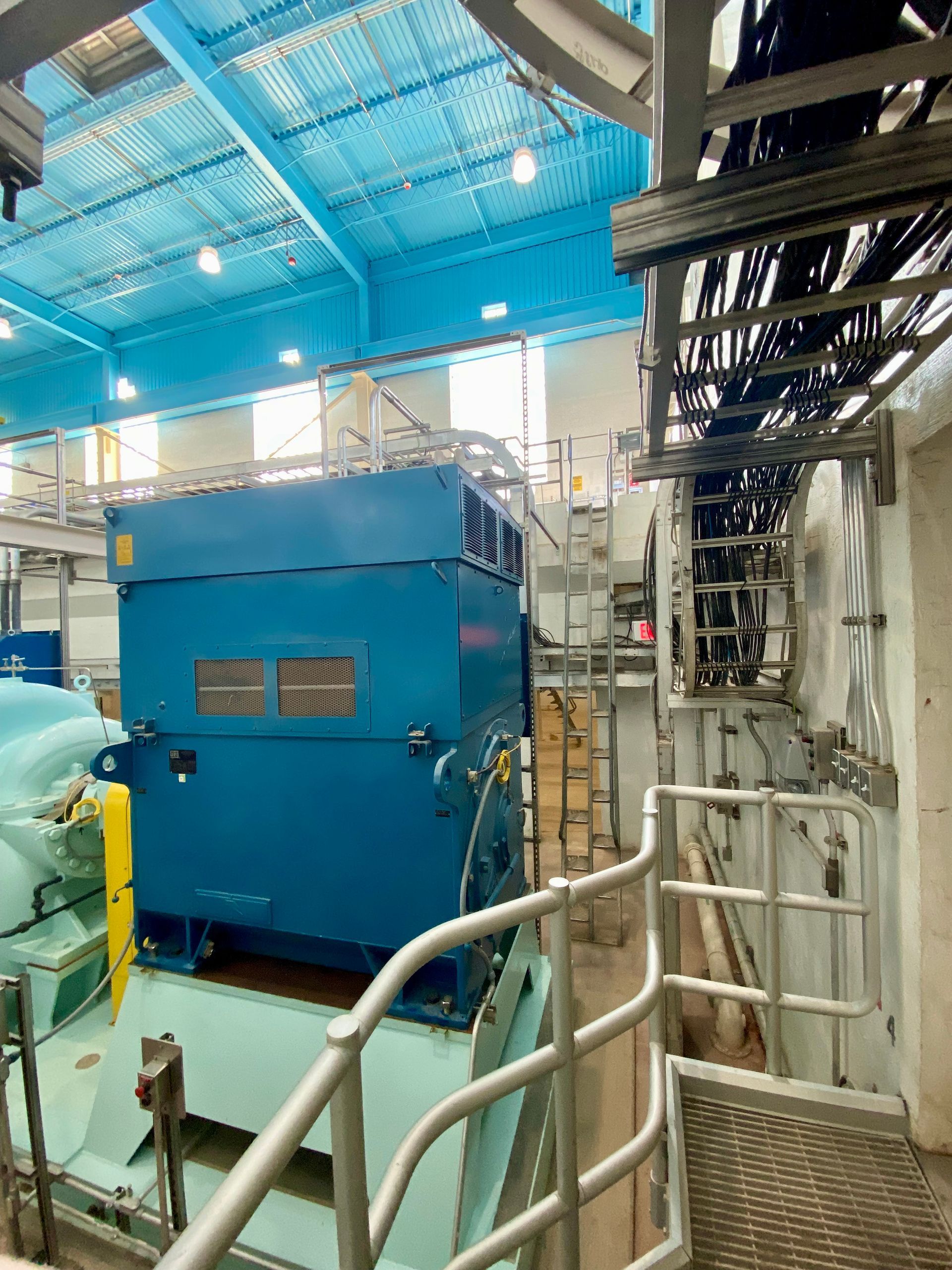 Blue industrial machine inside a factory, with metal railings and overhead piping.