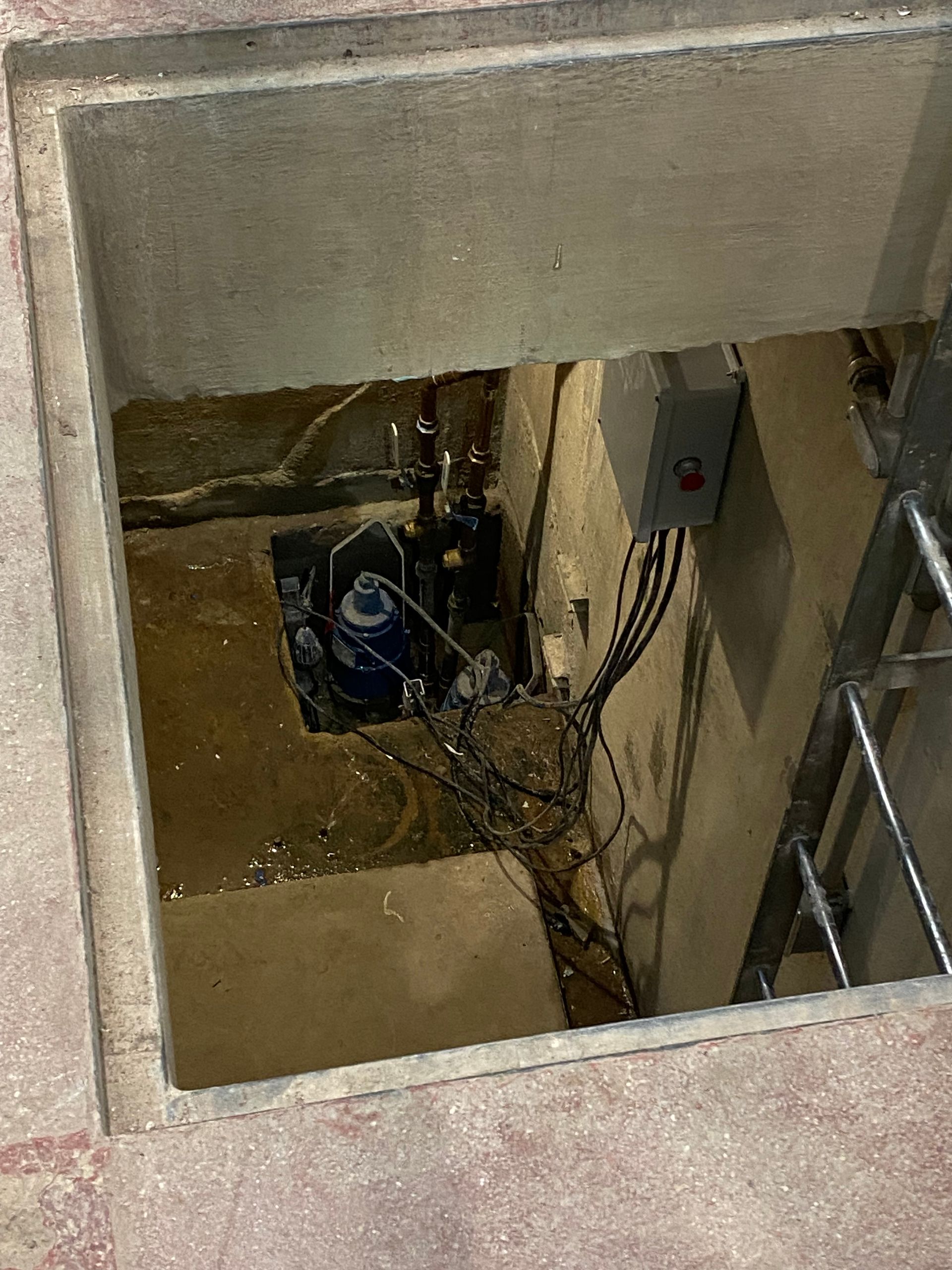 Open floor hatch revealing a small utility chamber with pipes, cables, and a pump