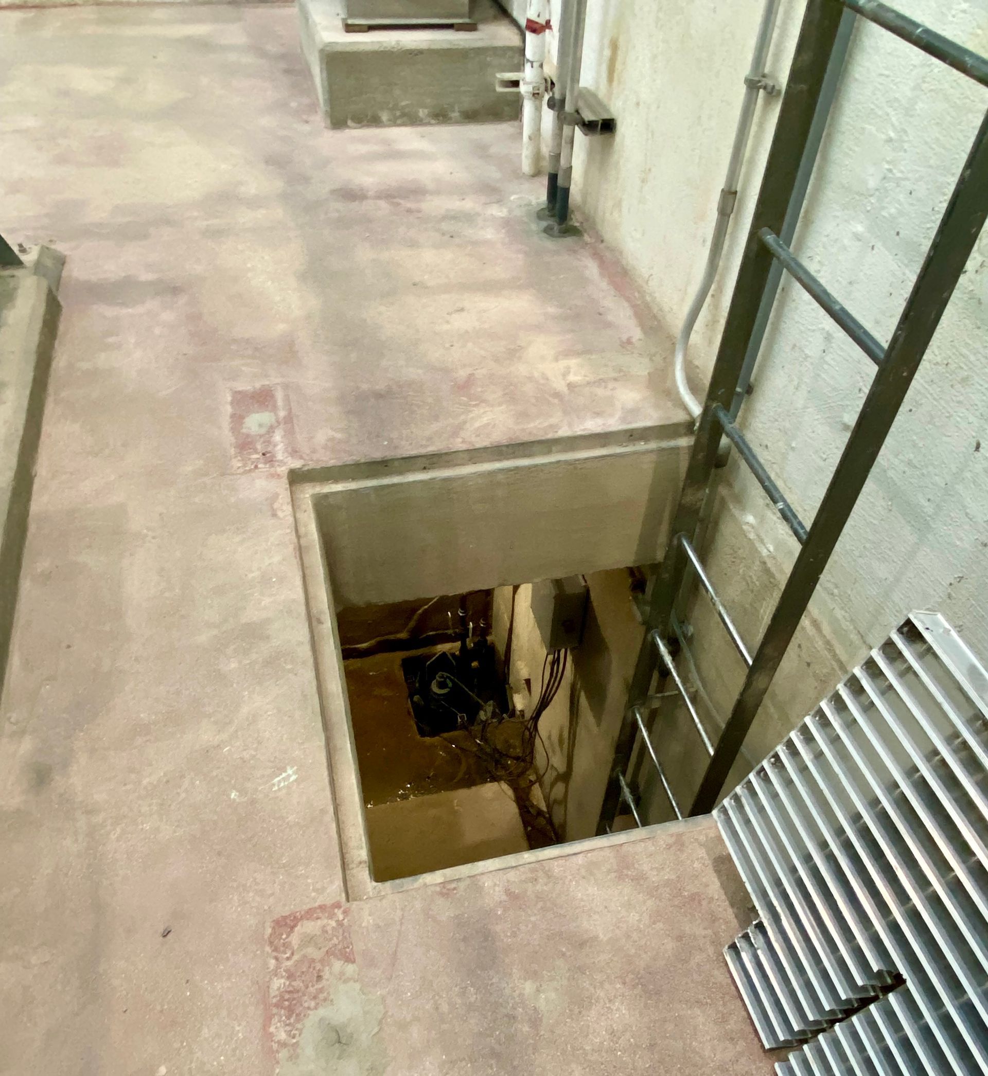 Open floor hatch with ladder descending into a utility shaft beside a concrete walkway.