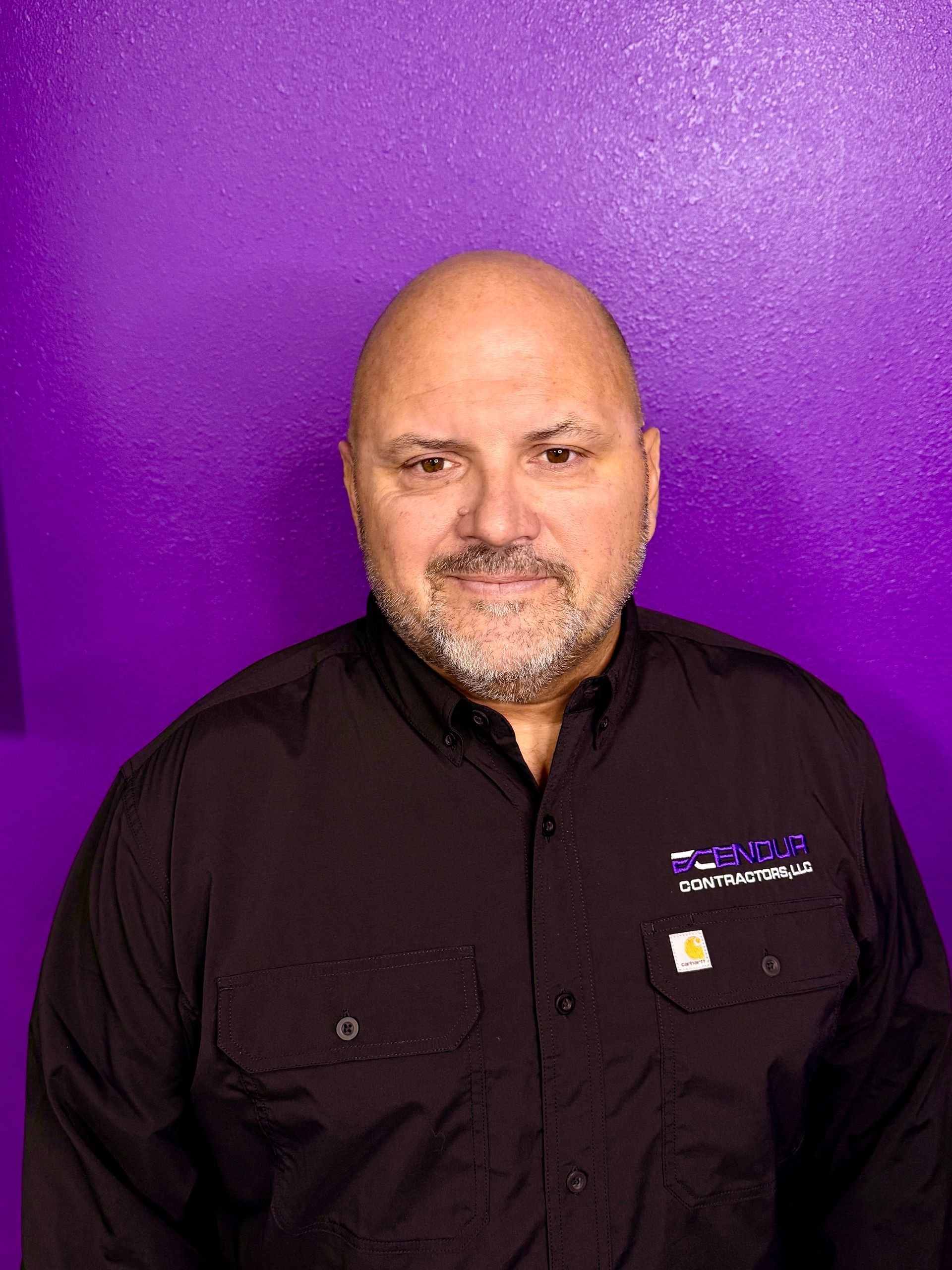 Bald man with a beard wearing a black shirt with a company logo, standing in front of a purple wall.