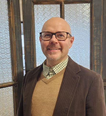 A bald man wearing glasses and a brown jacket is standing in front of a door.