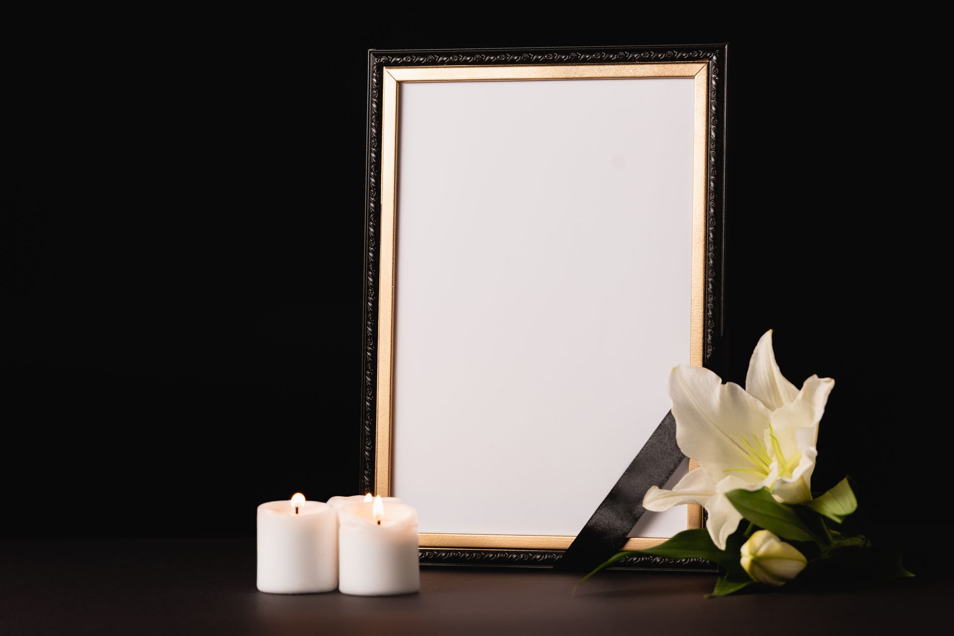 Cornice vuota con nastro nero, gigli bianchi e candele su una superficie scura.