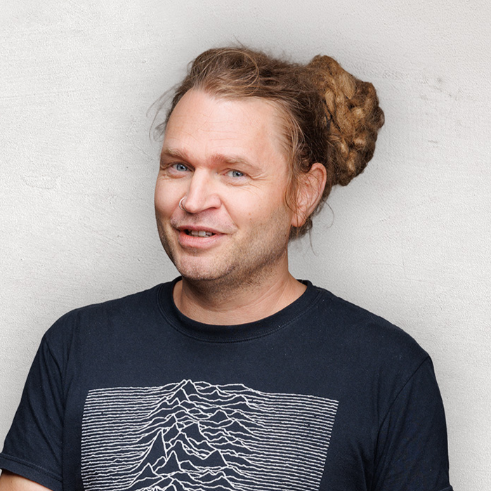 Mann mit lockigem Haar in einem dunklen T-Shirt mit Aufdruck, lächelt vor einer hellgrauen Wand.