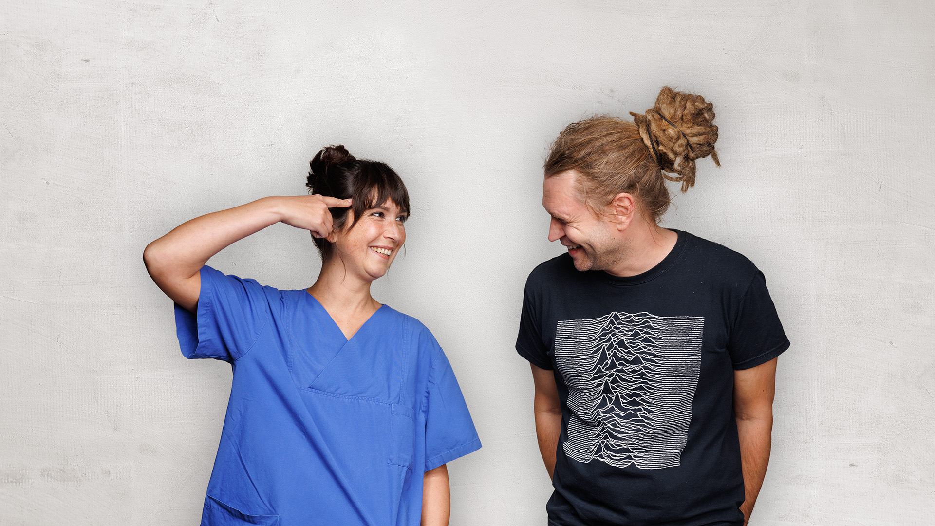 Zwei Personen lächeln sich an, eine in einem blauen Hemd, die andere in einem schwarzen T-Shirt mit Aufdruck, vor einer hellen Wand