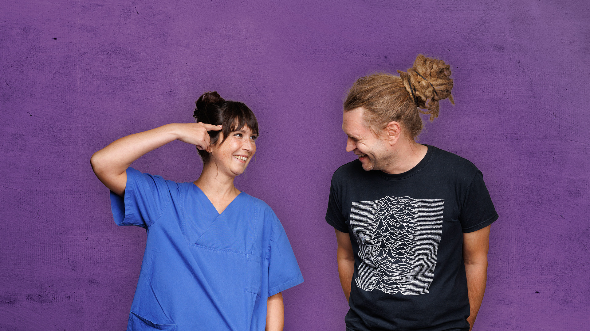 Zwei Personen lächeln und posieren vor einer lila Wand, eine in einem blauen Hemd und die andere in einem schwarzen T-Shirt mit Aufdruck.