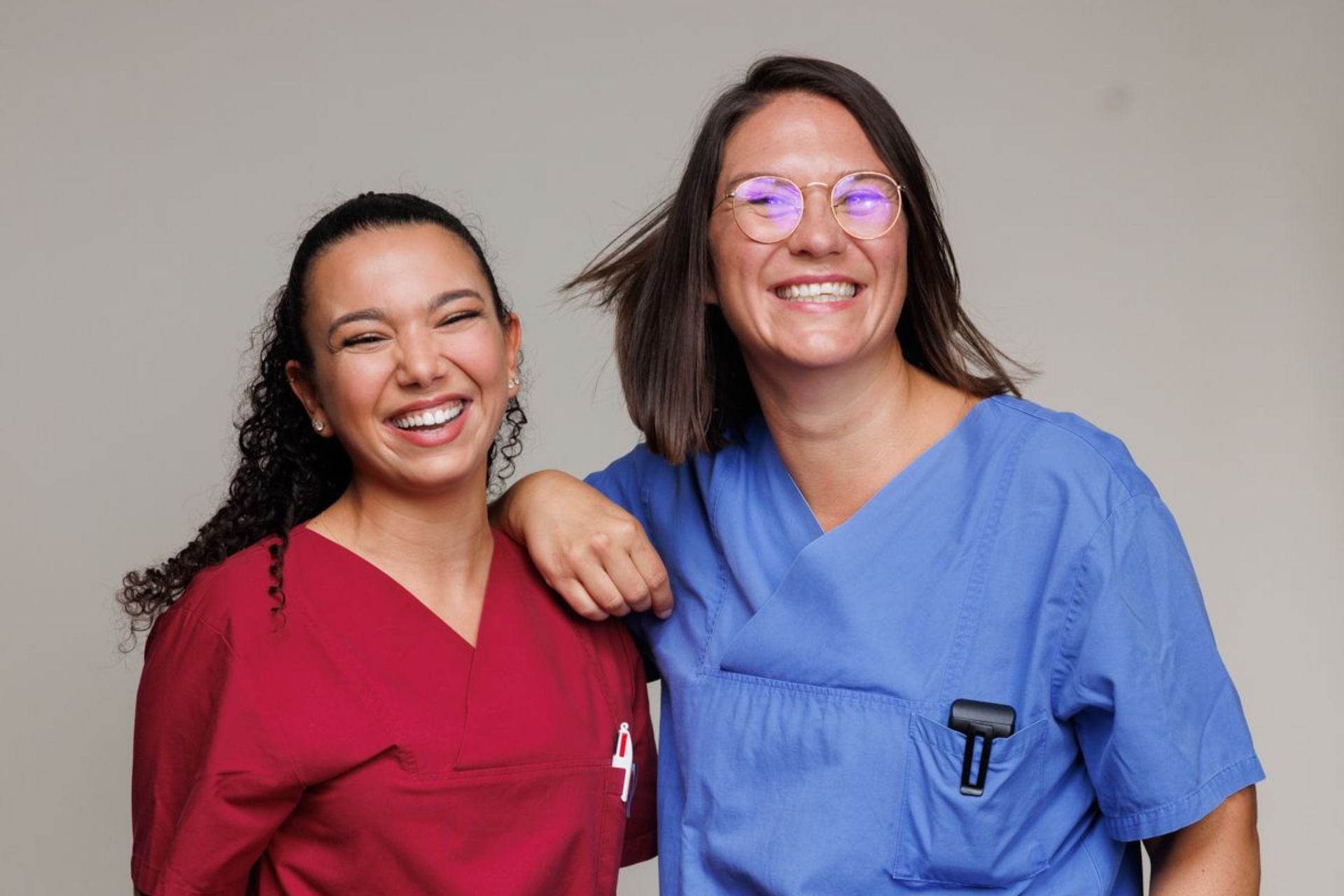 Zwei lächelnde Mitarbeiterinnen des Gesundheitswesens in roter und blauer OP-Kleidung stehen nebeneinander.