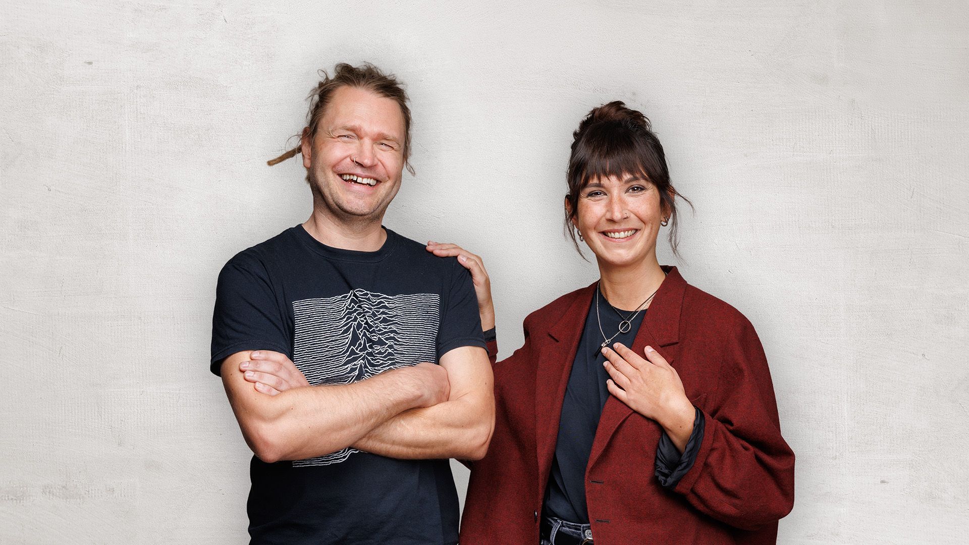 Zwei lächelnde Personen stehen vor einer hellen Wand, eine in einem schwarzen T-Shirt, die andere in einer roten Jacke.