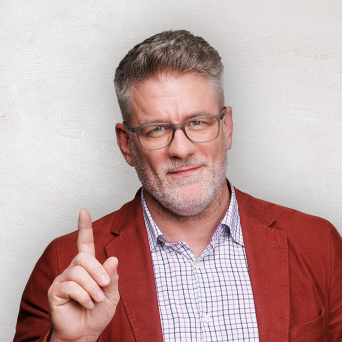 Ein Mann mit Brille und roter Jacke hebt vor einem hellgrauen Hintergrund einen Finger.