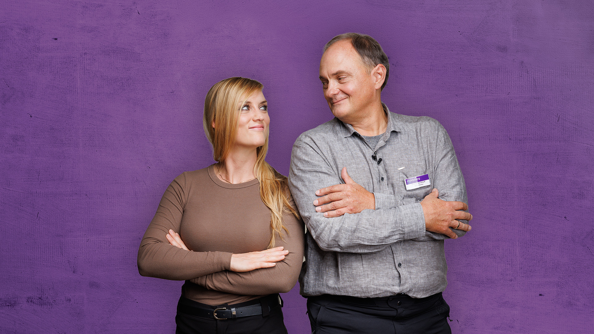 Zwei Personen stehen vor einem violetten Hintergrund, die Arme verschränkt, schauen sich an und lächeln.