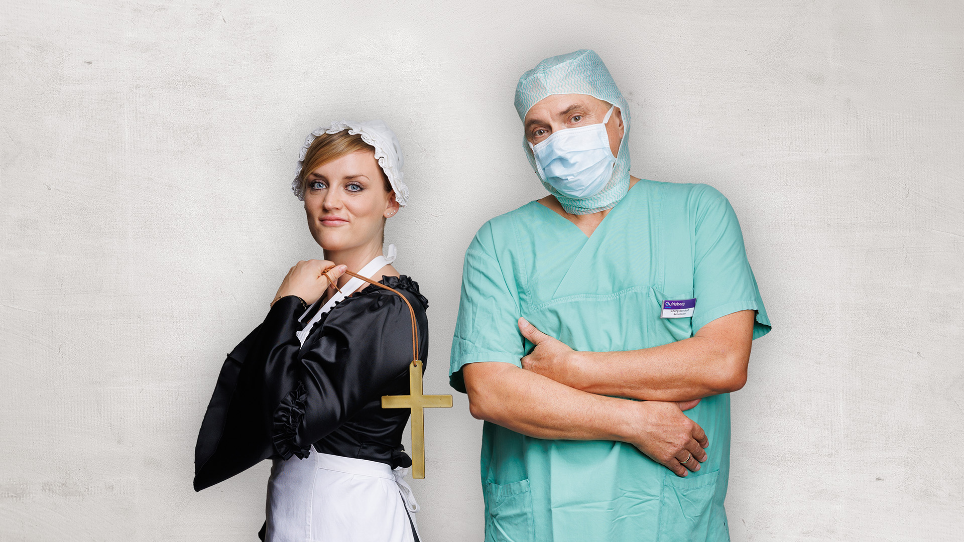 Zwei medizinische Fachkräfte in OP-Kleidung und Hauben stehen Rücken an Rücken, eine von ihnen mit Maske, die Arme verschränkt vor einer schlichten Wand.