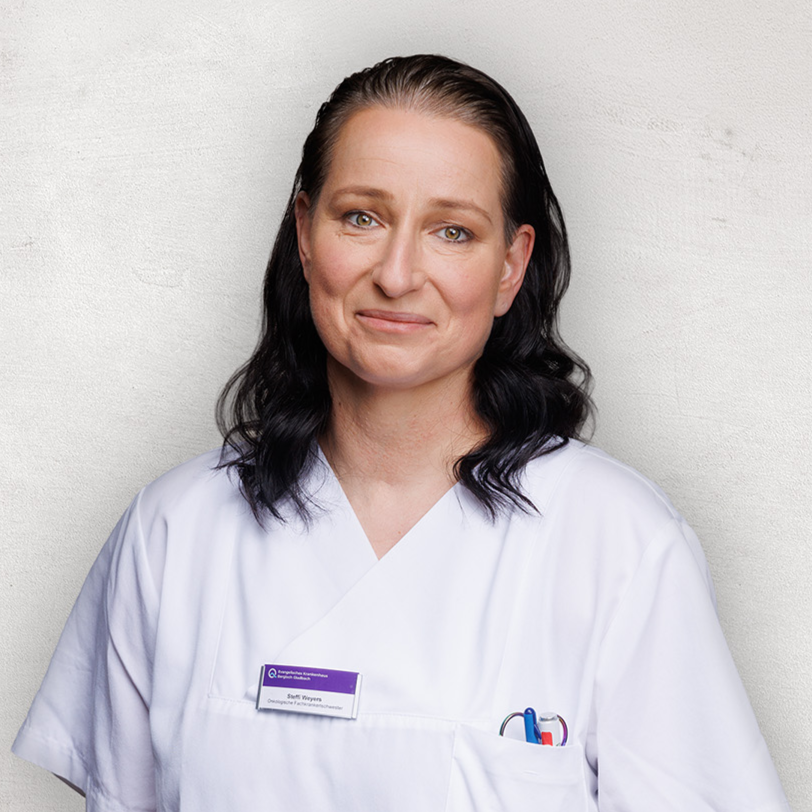 Medizinische Fachkraft in weißer Uniform, vor schlichtem Hintergrund, mit einem leichten Lächeln.