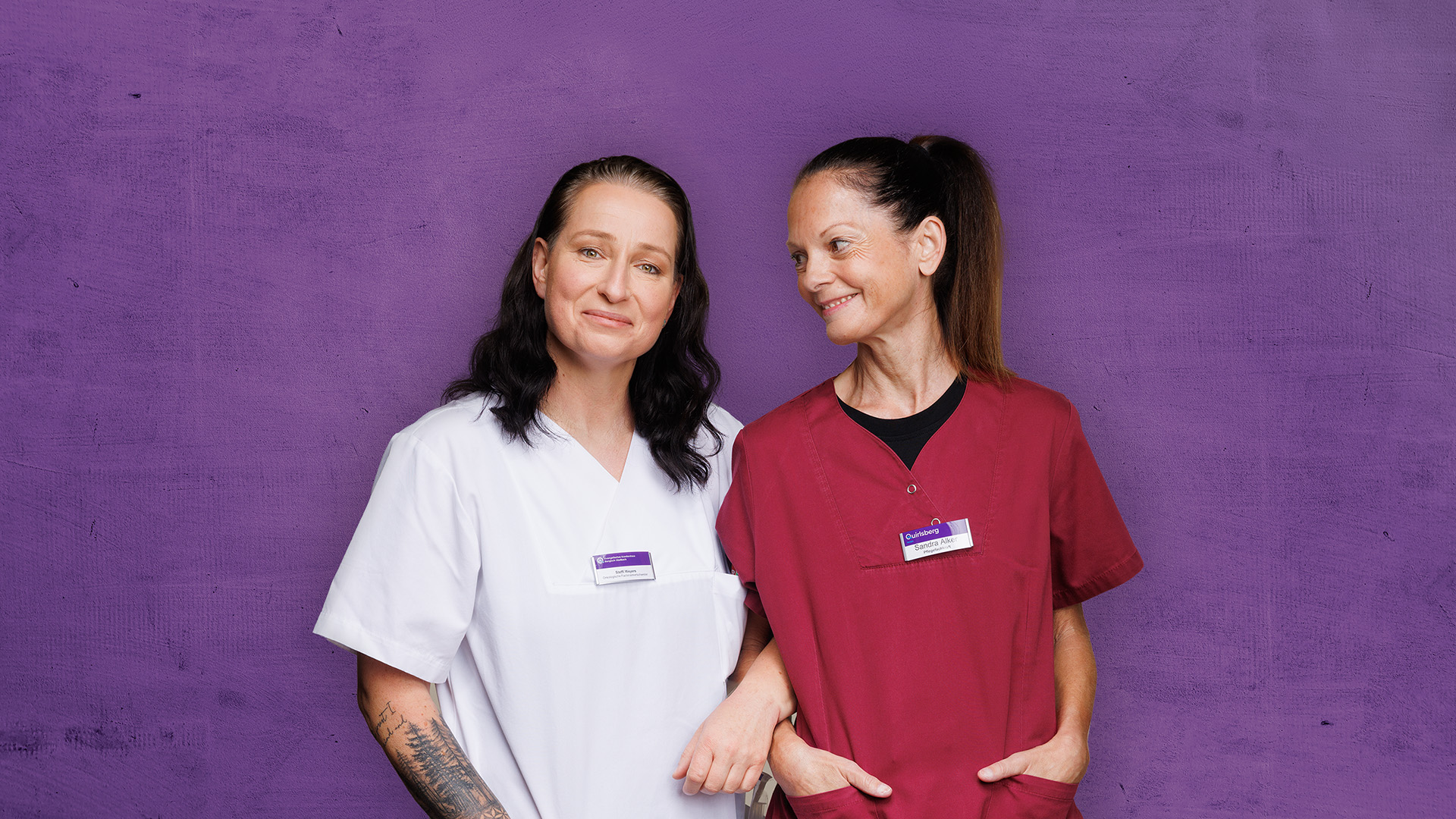 Zwei Personen stehen lächelnd vor einer violetten Wand und blicken einander an; sie tragen weiße und rote Hemden.