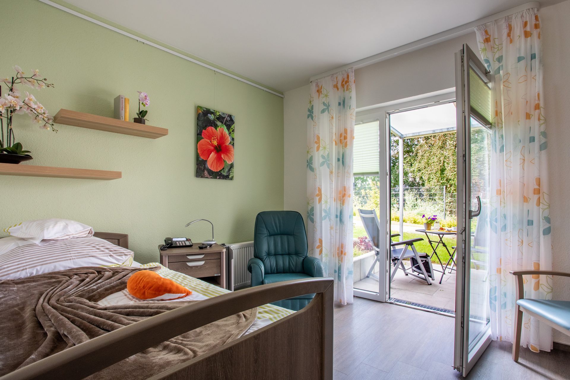 Helles Schlafzimmer mit grünen Wänden, Bett, Schreibtisch, Stuhl und offener Glastür zu einer sonnigen Terrasse.