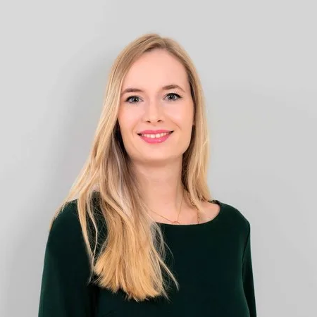Porträt einer lächelnden blonden Frau in einem dunkelgrünen Oberteil vor hellgrauem Hintergrund