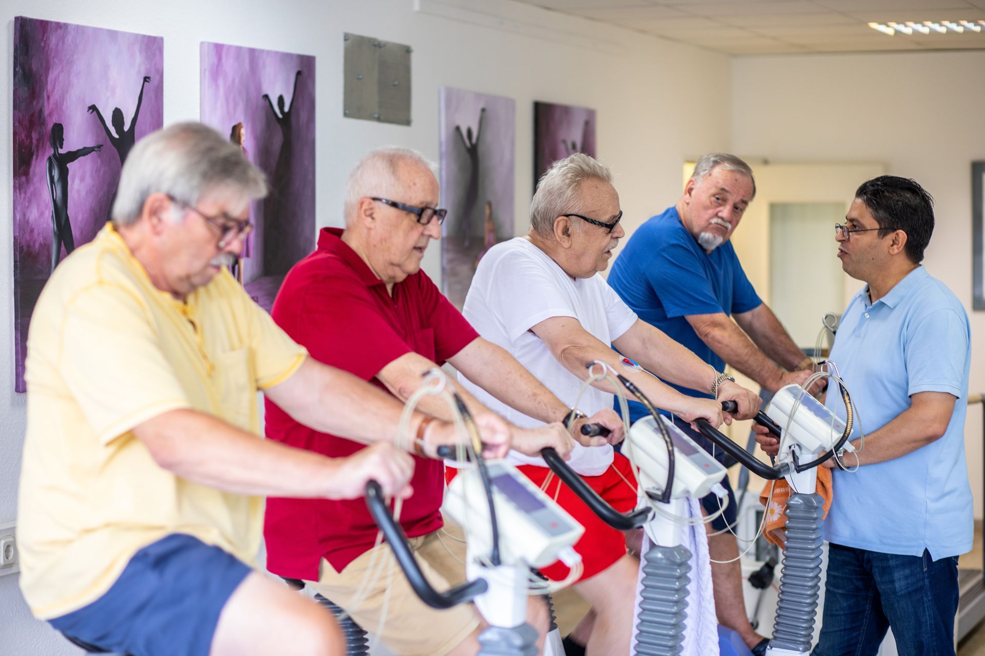 Vier Menschen trainieren in einem hellen Fitnessstudio und fahren Ergometer.