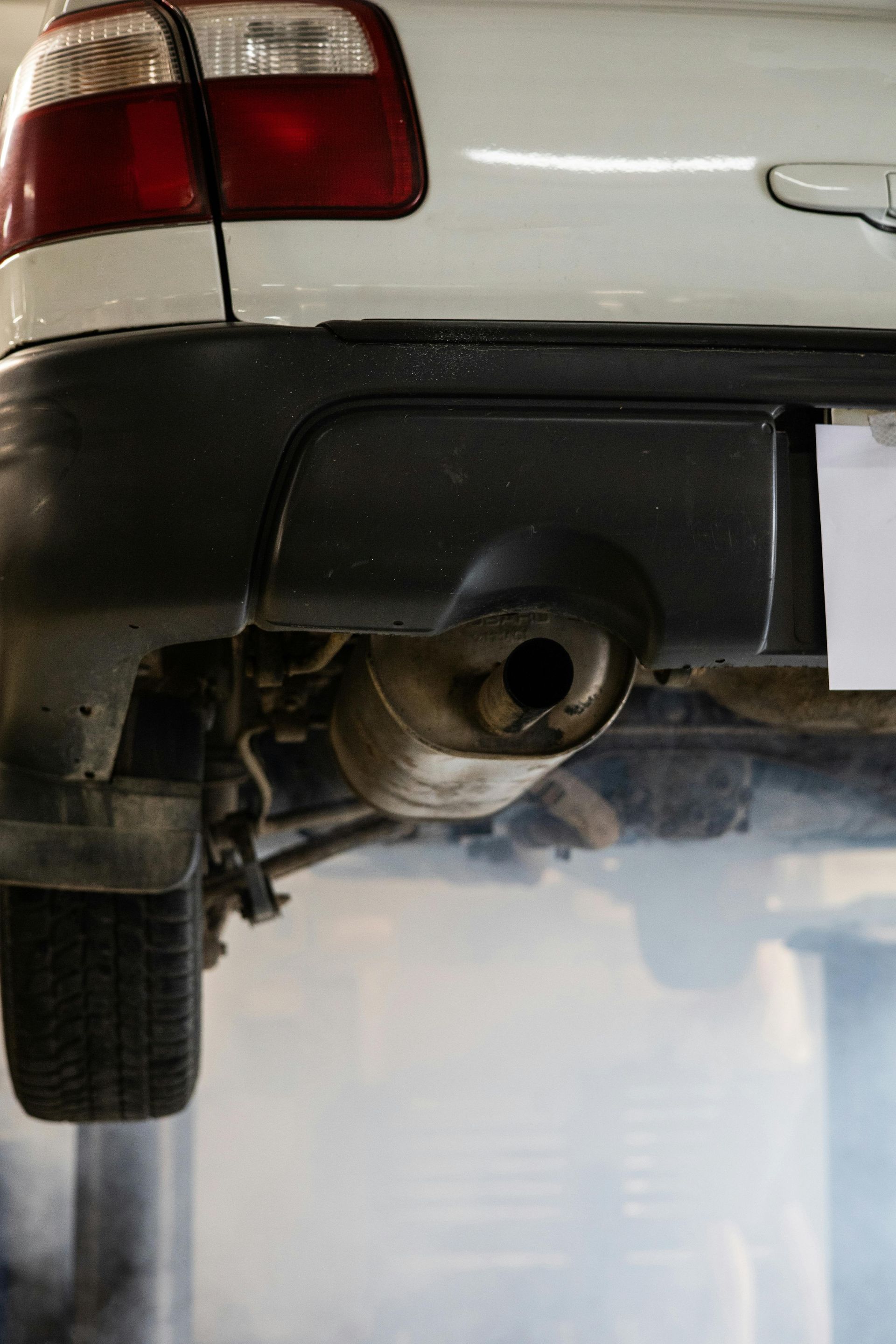 The rear of a white vehicle is raised on a lift, with thick white exhaust smoke billowing from its tailpipe.
