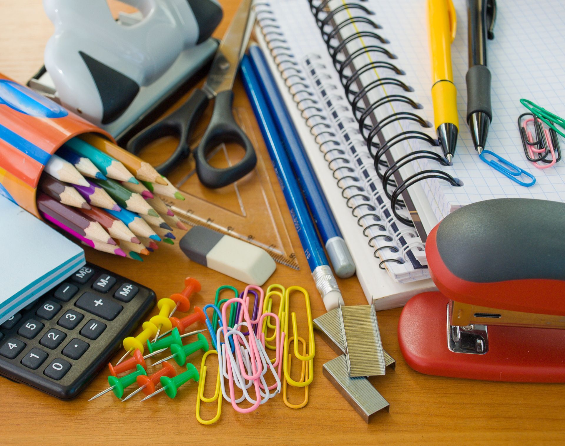 Útiles escolares variados, incluidos lápices, cuadernos, tijeras, una calculadora y chinchetas, dispuestos sobre una superficie de madera.