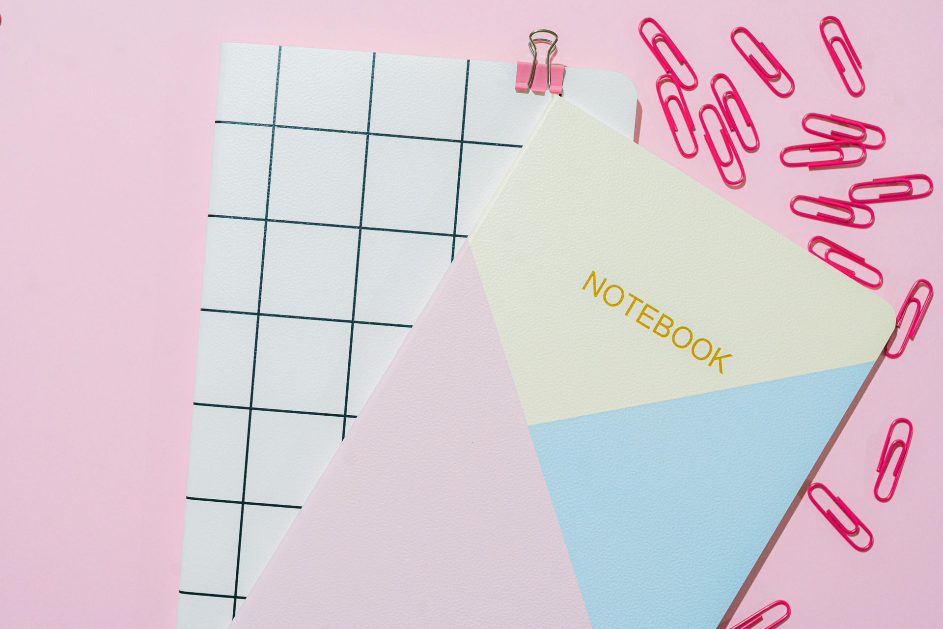 Escritorio rosa con papel cuadriculado, cuaderno, clips rosas y un cuaderno de color celeste y crema.