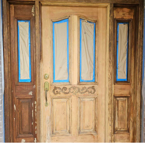 Wooden front door being refinished, with taped windows and stained side panels.