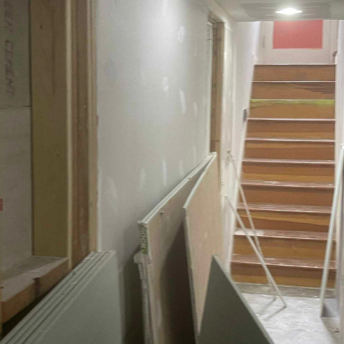 Construction site interior with drywall leaning against a wall, stairs and door frames visible.