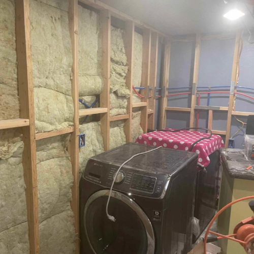 Laundry room under construction: insulation between wooden studs, washer, and utility lines visible.