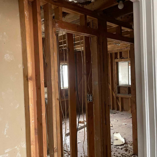 Interior view of a room under renovation. Studs and wiring exposed, debris on the floor, windows visible in the back.