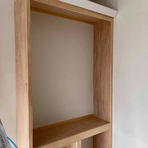 Wooden built-in shelving unit with one shelf; light wood, white wall backdrop.