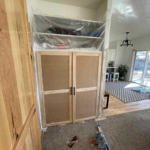 Built-in cabinet with doors and shelving, protected by plastic. Interior of a home with a rug and kitchen in the background.