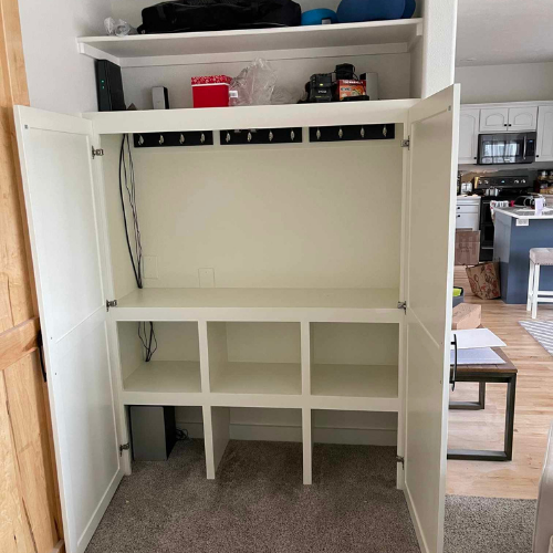 White storage cabinet, open, with shelves, hooks, and doors. In a room with a kitchen visible.