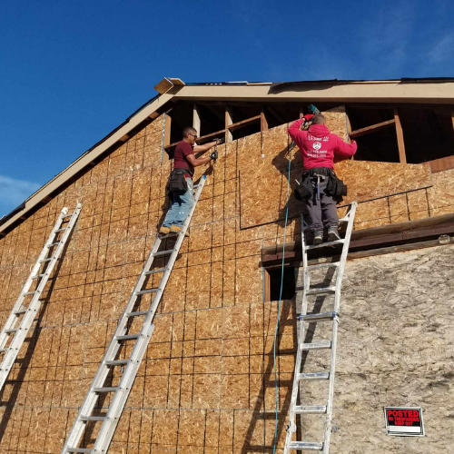 Two construction workers on ladders attaching wood panels to a building exterior under a blue sky.