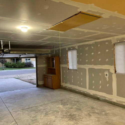 Garage interior with drywall work in progress; open garage door; cabinets on a wall.