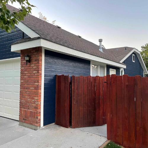 Exterior of a house with dark blue siding, a white garage door, red brick, and a wooden fence.