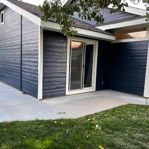 Dark blue house with white trim, sliding glass door, and concrete patio. Green lawn and a tree branch are visible.