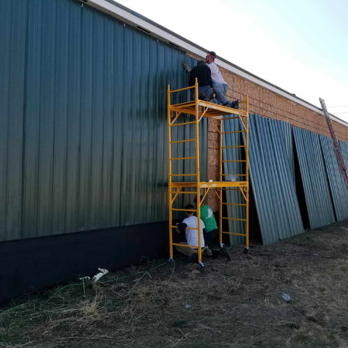 Workers installing green metal siding on a building, using scaffolding.