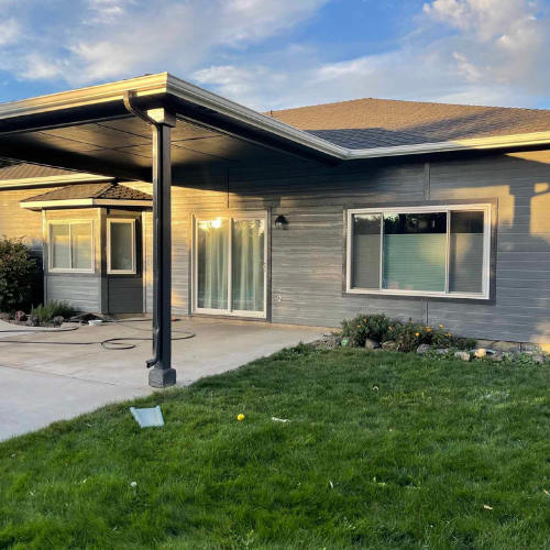 Gray house with covered carport, sliding glass door, and green lawn under a blue sky.