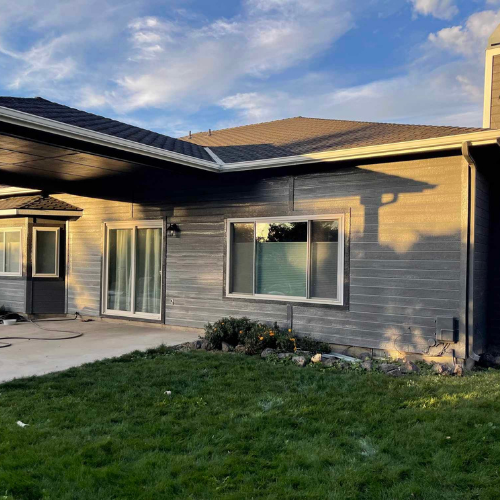Exterior view of a house with gray siding, a carport, and green lawn under a blue and cloudy sky.