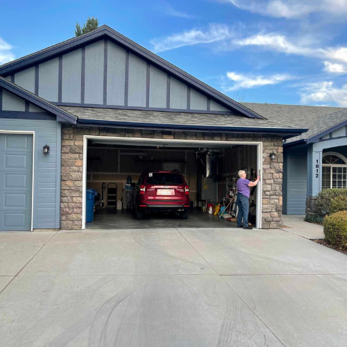 Garage with a red SUV, an open door, and a man standing inside. Blue house with a cloudy sky.