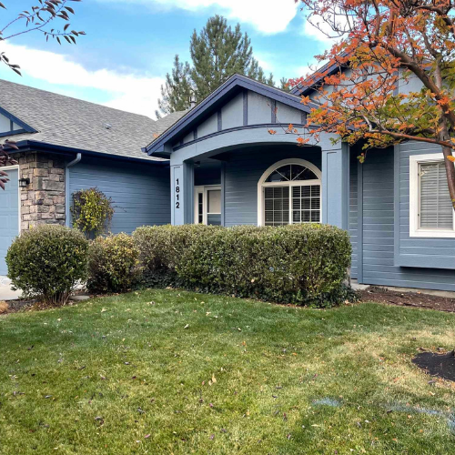 Blue house with gray roof, white arched window, green bushes, and a front lawn with fall foliage.