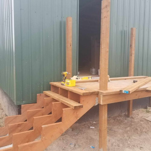 Wooden deck and staircase being built next to a green metal building, partially completed.