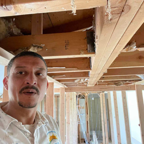 Man in a construction site looking at the camera. Exposed ceiling beams and wall studs visible.