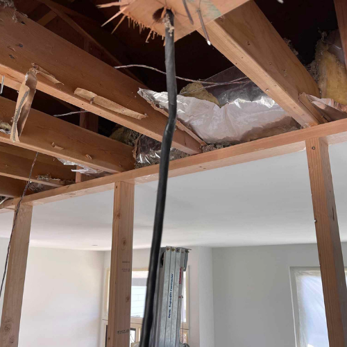 Interior view of a room under construction with exposed wooden beams, wiring, and insulation visible.