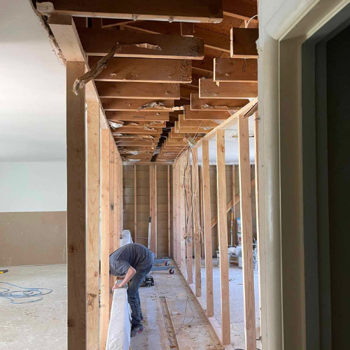 Interior undergoing renovation. A person works on exposed framing. Hallway, exposed ceiling joists.
