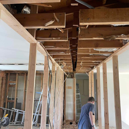 Interior during renovation, exposed wooden beams and framing. Man stands near doorway.