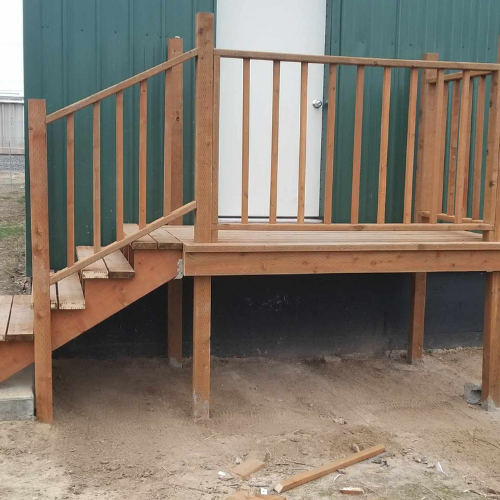 Wooden deck with stairs leading to a door. Handrails are present. Green building in the background.